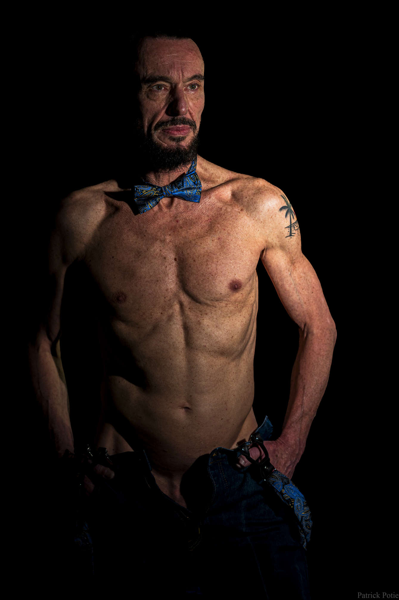 portrait d'un homme mince avec les veines et muscles marqués, portant un jeans, un nœud papillon et des bretelles.