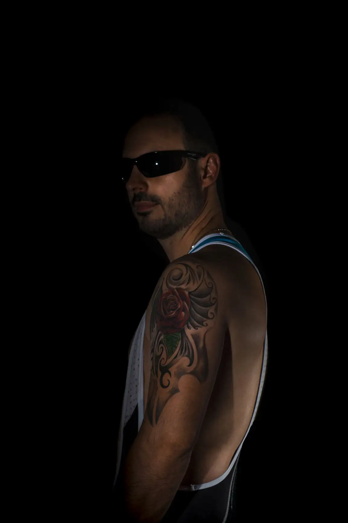 Un homme pose dans une lumière tamisée, montrant un tatouage de rose sur son bras et portant des lunettes de soleil.