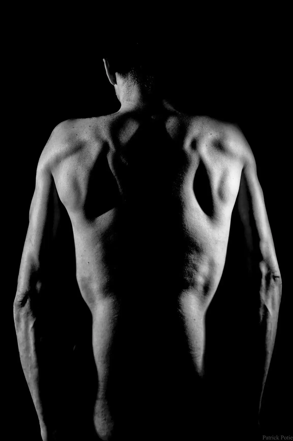 Un homme exhibe son dos et ses bras musclés, sa silhouette athlétique, sous un éclairage dramatique.