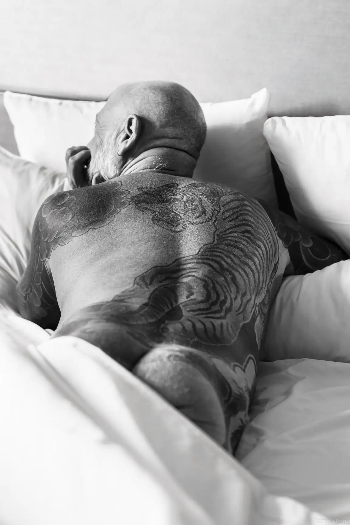 Un homme chauve se repose sur des coussins blancs, le dos couvert de tatouages ​​détaillés de tigres et de nuages.